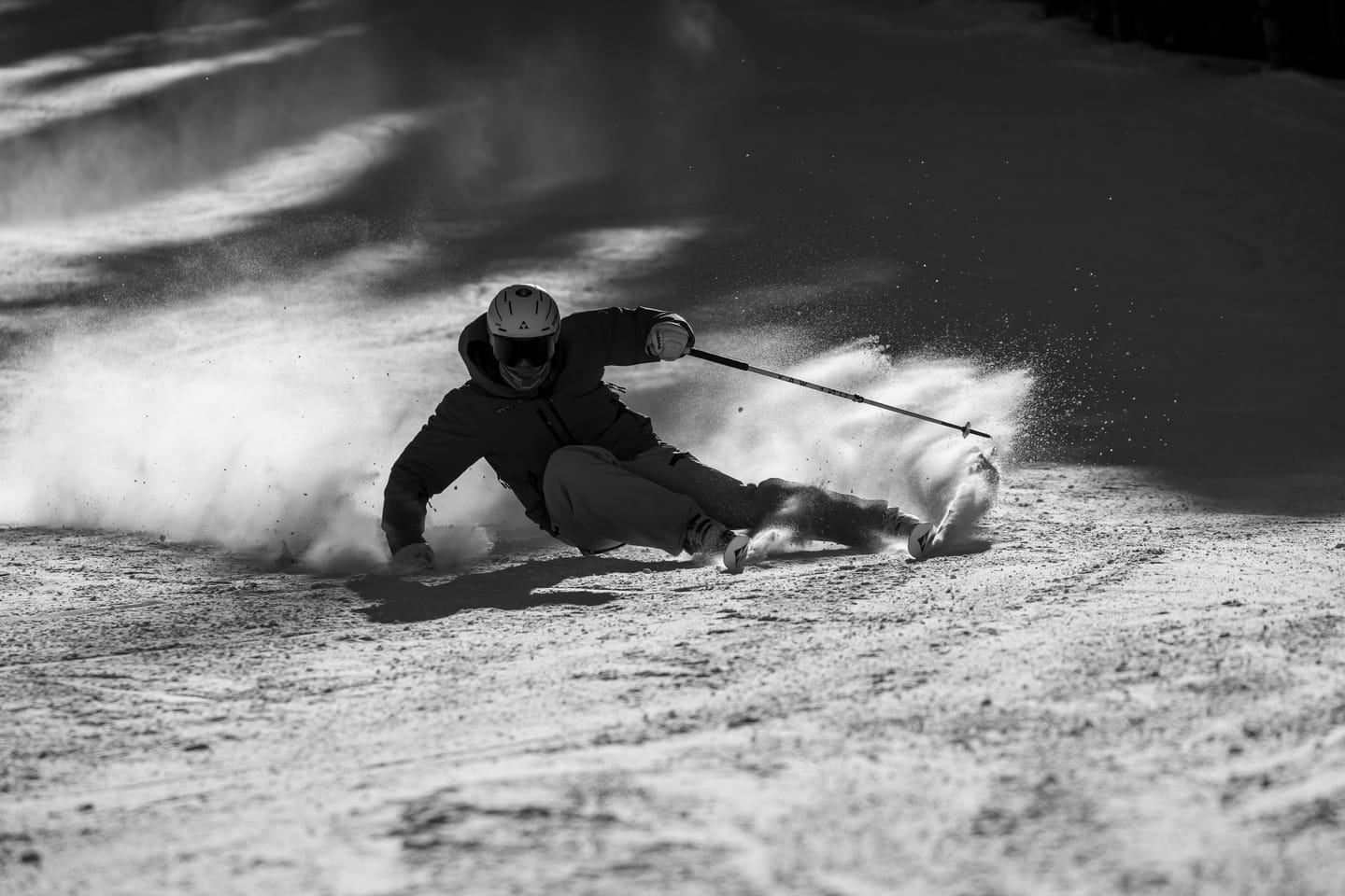 Black and white photo of Tomas Mical in a deep carved turn, snow spraying behind him
